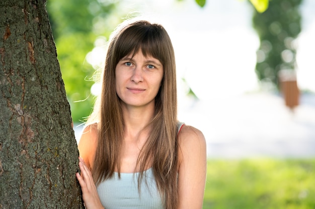 Porträt der hübschen jungen Frau mit den langen Haaren, die ordentlichen Baumstamm im Sommer stehen.