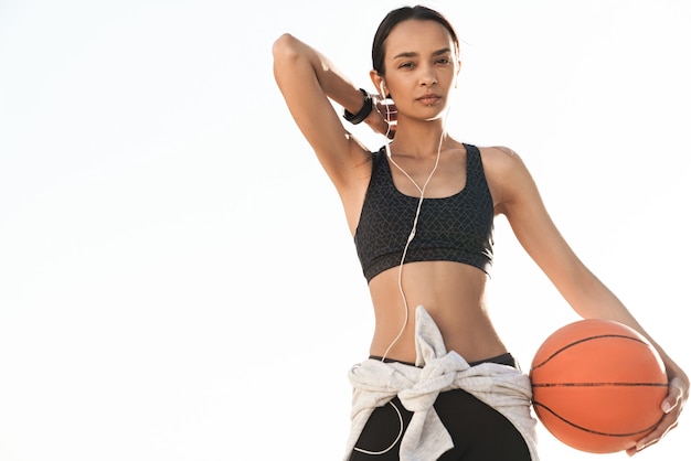 Porträt der hübschen schönen starken Sportfrau draußen am Strand, die hörende Musik mit Kopfhörern aufwirft, die Basketball halten.