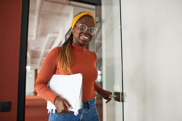 Porträt der jungen afrikanischen Geschäftsfrau, die an der Kamera lächelt, während sie beim Treffen ins Büro geht