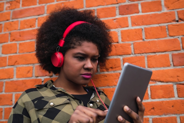 Porträt der jungen afroamerikanischen Frau, die ihr digitales Tablett mit roten Kopfhörern im Freien verwendet. Technologiekonzept.