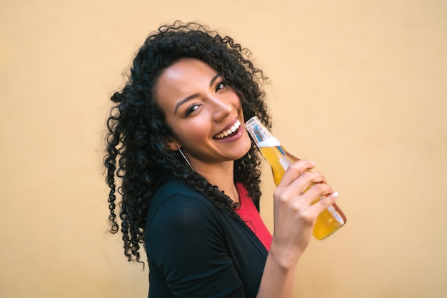 Porträt der jungen afroamerikanischen lateinamerikanischen Frau, die eine Flasche Bier genießt und trinkt, gegen gelben Hintergrund. Lifestyle-Konzept.