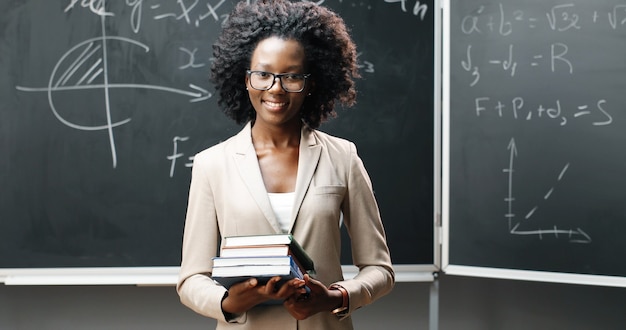 Porträt der jungen afroamerikanischen Lehrerin in den Gläsern, die Kamera im Klassenzimmer betrachten und Lehrbücher halten. Tafel mit Formeln auf Hintergrund. Schulkonzept. Bücher in Frauenhänden.