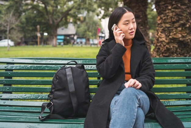 Porträt der jungen asiatischen Frau, die am Telefon spricht, während sie auf einer Bank im Park sitzt. Kommunikationskonzept.