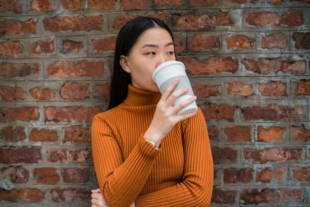 Porträt der jungen asiatischen Frau, die eine Tasse Kaffee gegen Mauer trinkt. Stadtkonzept.