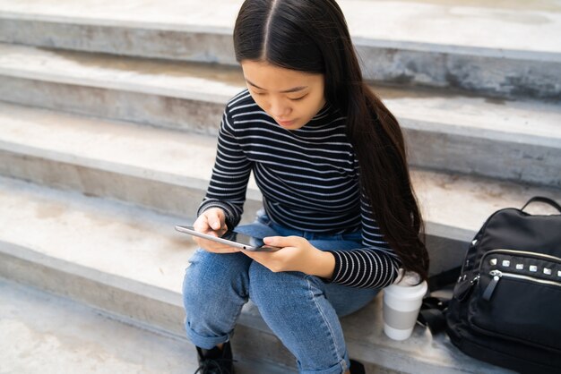 Porträt der jungen asiatischen Frau, die ihr digitales Tablett beim Sitzen an der Treppe im Freien verwendet. Technologiekonzept.