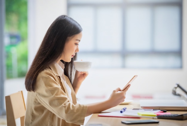 Porträt der jungen asiatischen Geschäftsfrau, die drinnen im Café sitzt, das Kaffee mit Smartphone trinkt. Geschäftserfolgskonzept.