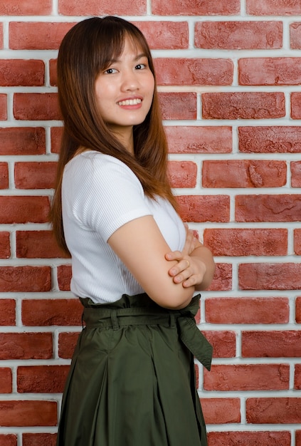 Porträt der jungen attraktiven asiatischen Frau im stehenden Arm des weißen T-Shirts und des grünen Rockes gekreuzt und lächelnd mit orange Backsteinmauerhintergrund.