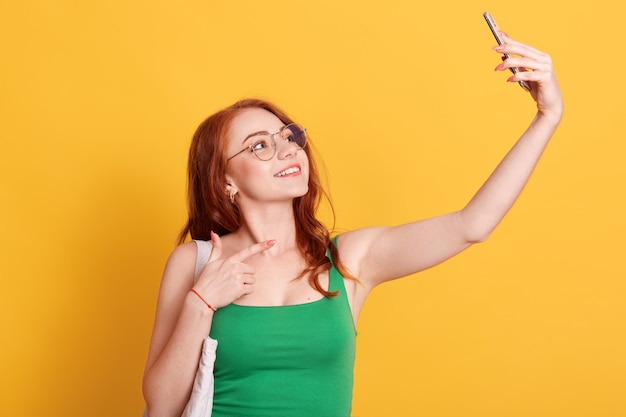 Porträt der jungen attraktiven Frau, die Selfie-Foto mit Smartphone lokalisiert über gelbem Hintergrund macht, rothaarige Frau in grünem T-Shirt zeigt auf Telefonkamera mit Zeigefinger.