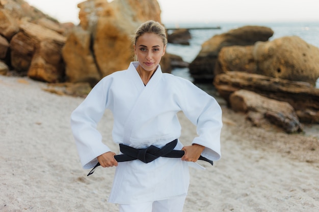 Porträt der jungen attraktiven Kampfkunstmeisterin im weißen Kimono mit schwarzem Gürtel am wilden Strand