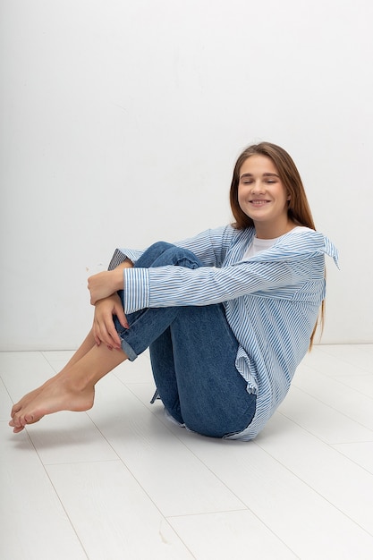 Porträt der jungen attraktiven kaukasischen Frau mit dem langen braunen Haar im T-Shirt und in den blauen Jeans lokalisiert auf weißem Hintergrund. Skinny hübsche Frau sitzt auf dem Boden im Studio. Modelltests der schönen Dame