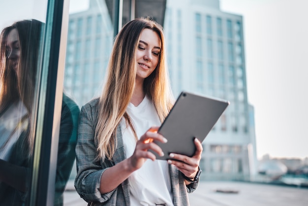 Porträt der jungen attraktiven lässig gekleideten Geschäftsfrau mit einer Tablette in den Händen, die vor dem High-Tech-Glasgebäude des Handelszentrums aufwirft