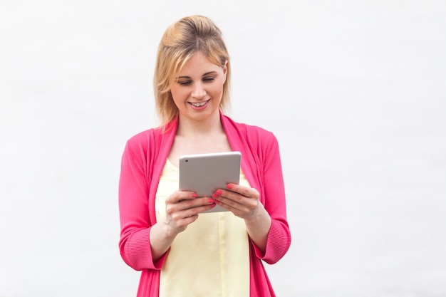 Porträt der jungen Frau der schönen erfolgreichen Geschäftsfrau in der rosa Bluse, die interessante Mitteilung auf ihrem Tablet mit glücklichem Gesicht steht, verwendet und liest. Innen, isoliert, Studioaufnahme, weißer Hintergrund