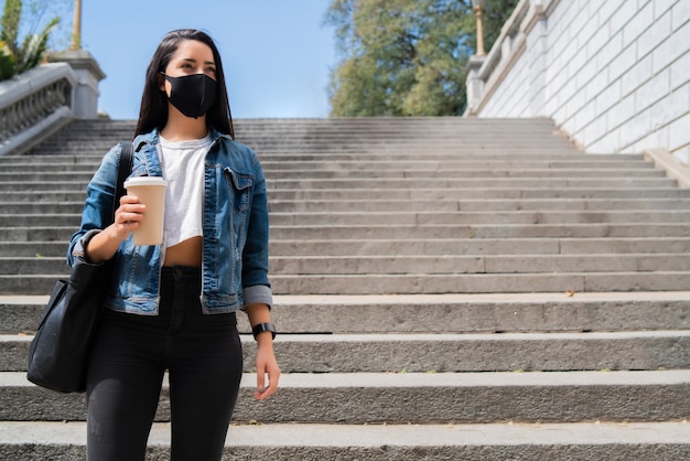 Porträt der jungen Frau, die Gesichtsmaske trägt und eine Tasse Kaffee hält, während draußen steht. Stadtkonzept. Neues normales Lifestyle-Konzept.