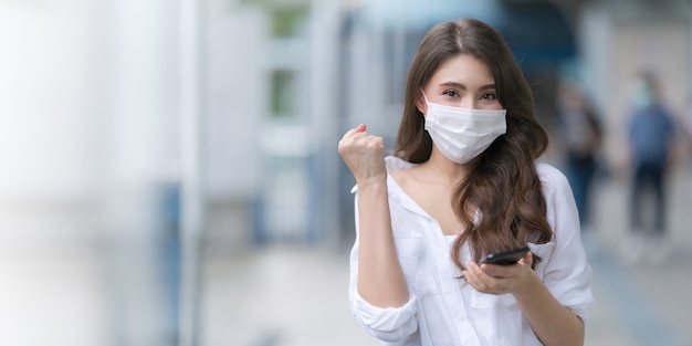 Porträt der jungen Frau, die medizinische Gesichtsmaskenschutz mit einem Telefon trägt, geht in eine Stadt