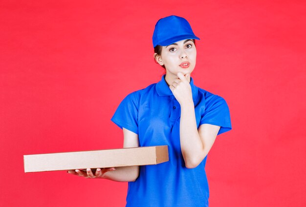 Porträt der jungen Frau in der blauen Ausstattung, die mit Pappschachtel über roter Wand aufwirft.