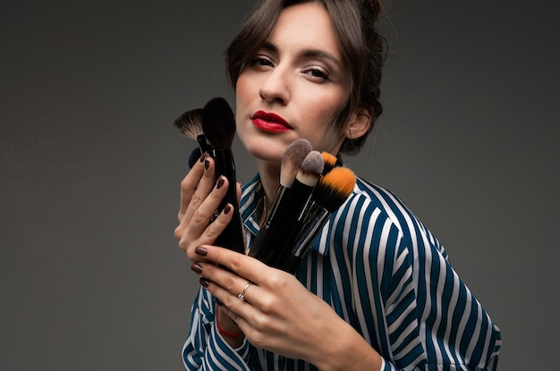 Porträt der jungen Frau in der gestreiften Bluse, die Make-upbürsten in ihren Händen lokalisiert hält