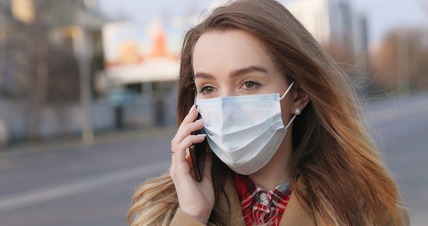 Porträt der jungen Frau in der medizinischen Gesichtsmaske, die auf einem Smartphone auf der Straße spricht. Coronavirus-Quarantäne. Pandemiekonzept, Coronavirus-Epidemie.