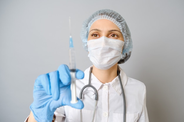 Porträt der jungen Frau in der medizinischen Uniform, in der Schutzmaske und in der Kappe mit der medizinischen Spritze in der Hand. Auf grauem Hintergrund isoliert