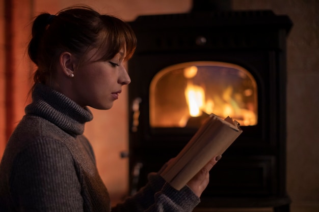 Porträt der jungen Frau in einem warmen Pullover, der ein Buch zu Hause am Kamin liest