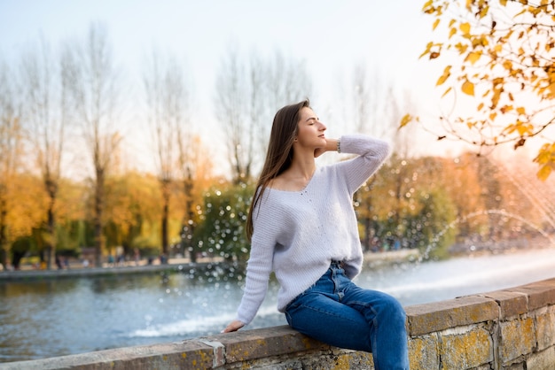 Porträt der jungen Frau in Freizeitkleidung, die draußen nahe Brunnen aufwirft. Herbst in der Stadt