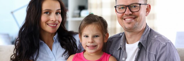 Porträt der jungen fröhlichen Leute, die auf Sofa sitzen. Vater in Brille umarmt süße hübsche Tochter. Schöne schöne Mutter, die Kamera mit Glück betrachtet. Elternschafts- und Kindheitskonzept