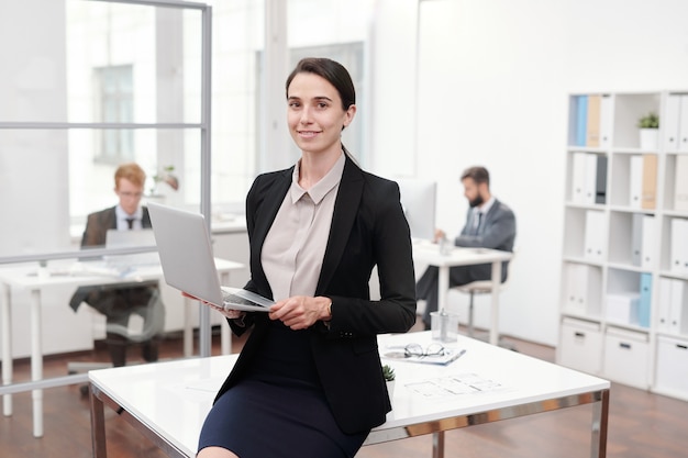 Porträt der jungen Geschäftsfrau, die Laptop hält, während auf Schreibtisch im Büro, Kopierraum gelehnt