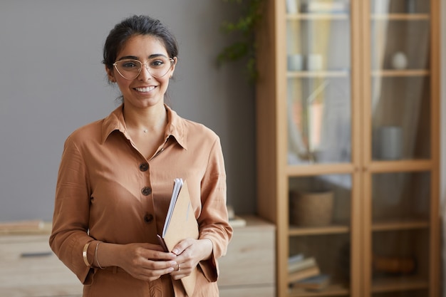 Porträt der jungen Geschäftsfrau in den Brillen und in der Bluse, die Ordner hält und vorne im Büro lächelt
