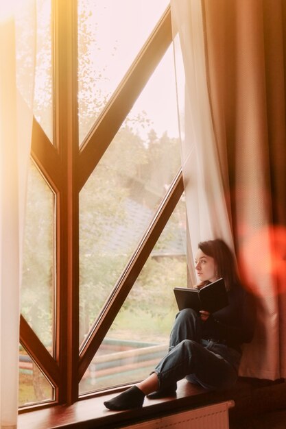 Porträt der jungen Geschäftsfrau mit Notebook und Telefon am Fenster des Wohnzimmers im Landhaus. Workaholic-Frau in Freizeitkleidung zu Hause, die im Urlaub arbeitet. Kreative Inspiration und Business