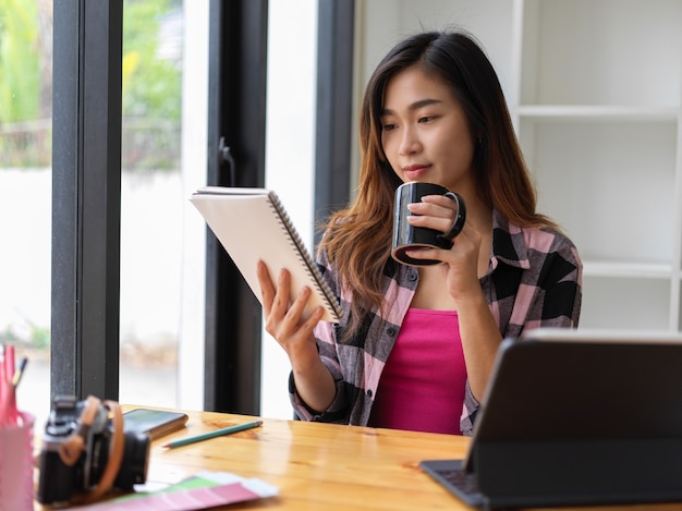 Porträt der jungen hübschen Frau, die Kaffeetasse und digitale Tablette beim Entspannen im Café hält