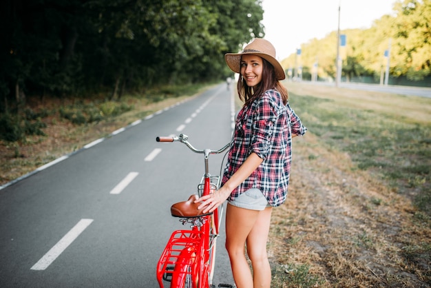 Porträt der jungen hübschen Frau im Hut mit dem roten Weinlesefahrrad, grüner Sommerpark. Radfahren im Freien. Mädchen auf Retro-Zyklus