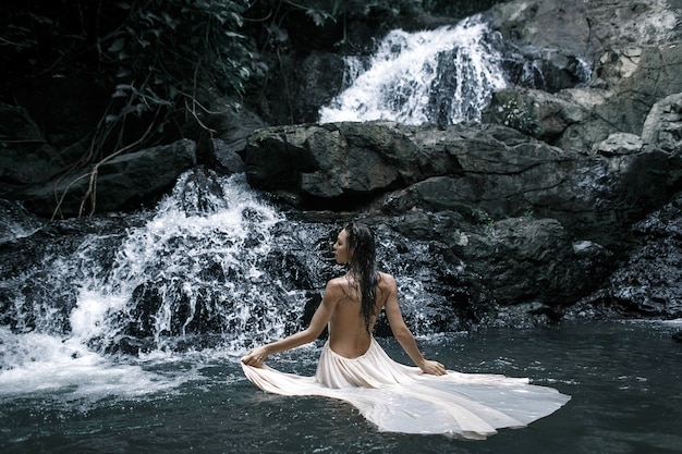Porträt der jungen kaukasischen Frau, die vor einem Wasserfall in einem weißen Abendkleid genießt. Rückansicht