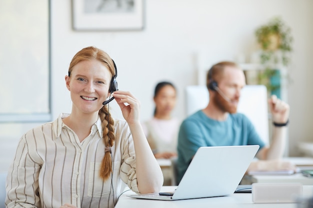 Porträt der jungen Kundendienstfrau in den Kopfhörern, die am Tisch mit Laptop sitzen und im Büro lächeln