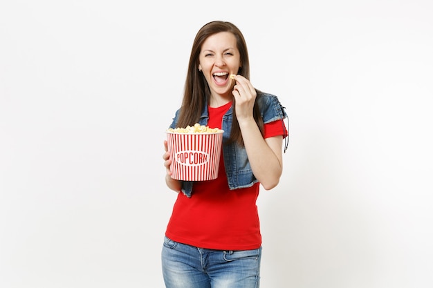 Porträt der jungen lachenden lustigen schönen Brunettefrau in der zufälligen Kleidung, die Filmfilm ansieht, Popcorn vom Eimer hält und isst, der auf weißem Hintergrund lokalisiert wird. Emotionen im Kinokonzept.