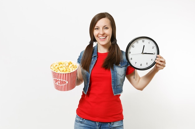 Porträt der jungen lächelnden attraktiven brünetten Frau in der Freizeitkleidung, die Filmfilm ansieht, Eimer Popcorn und runden Wecker einzeln auf weißem Hintergrund haltend. Emotionen im Kinokonzept.