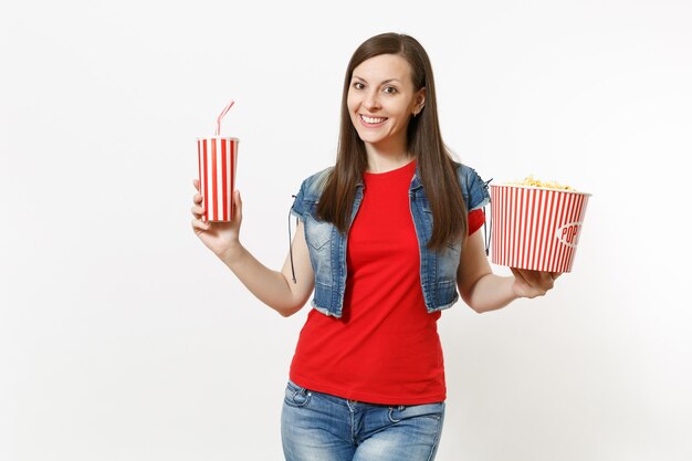 Porträt der jungen lächelnden attraktiven Frau in der Freizeitkleidung, die Filmfilm ansieht, Eimer Popcorn und Plastikbecher Soda oder Cola lokalisiert auf weißem Hintergrund haltend. Emotionen im Kinokonzept.