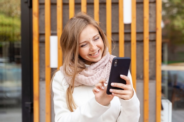 Porträt der jungen lächelnden Frau im weißen Mantel mit dem Schal, der den Smartphone berührt