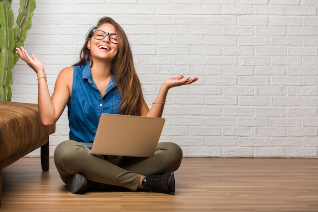 Porträt der jungen lateinischen Frau, die auf dem Boden sitzt, lachend und Spaß habend, entspannt und nett ist, fühlt sich überzeugt und erfolgreich. Einen Laptop halten.