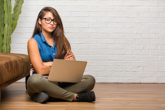 Porträt der jungen lateinischen frau, die auf dem boden sitzt, seine arme kreuzend, ernst und imposant, glaubend und leistung zeigend. einen laptop halten.