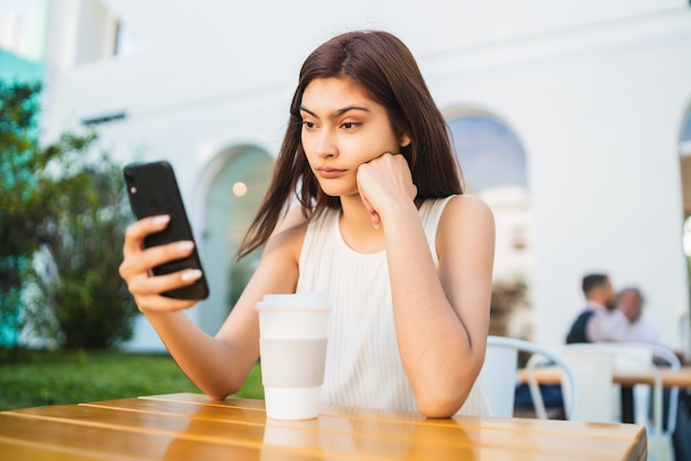 Porträt der jungen lateinischen Frau, die ihr Handy beim Sitzen im Coffeeshop im Freien verwendet