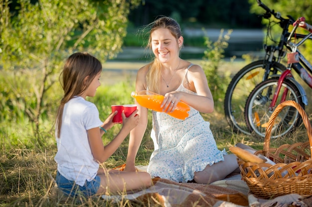 Porträt der jungen Mutter, die beim Picknick im Park Saft aus der Flasche in die Tasse der Tochter gießt