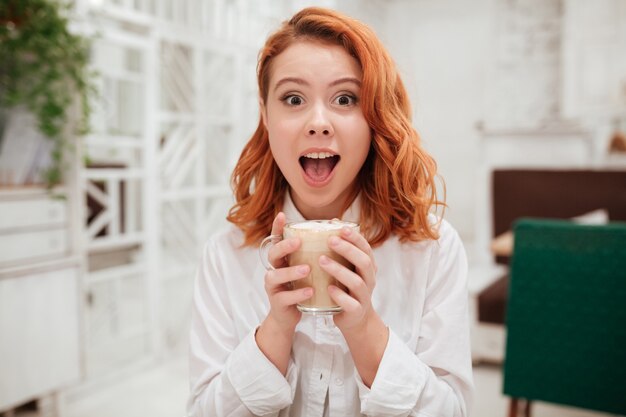 Porträt der jungen Rothaarigen überraschte glückliche Frau, die Kaffee im Café trinkt.