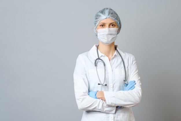 Porträt der jungen Ärztin in der medizinischen Schutzmaske auf ihrem Gesicht und Kappe auf ihrem Kopf. Auf grauer Wand isoliert