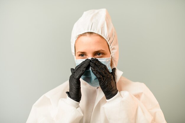 Porträt der jungen Sanitäterin im weißen Schutzanzug mit Kapuze, die chirurgische Maske auf Gesicht korrigiert.