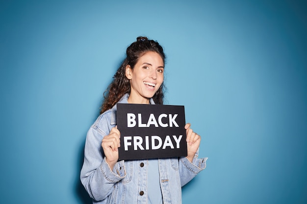 Porträt der jungen schönen Frau, die Black Friday Plakat hält und gegen den blauen Hintergrund lächelt
