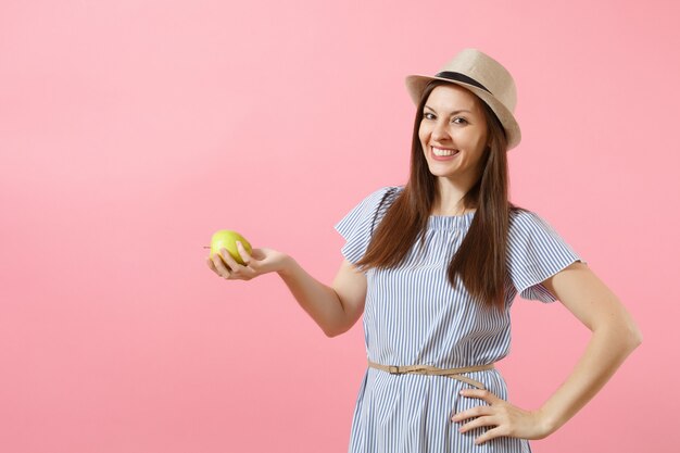 Porträt der jungen schönen Frau im blauen Kleid, Sommerstrohhutholding, grüne frische Apfelfrucht einzeln auf rosafarbenem Hintergrund essend. Gesunder Lebensstil, Menschen, Konzept der aufrichtigen Emotionen. Platz kopieren.