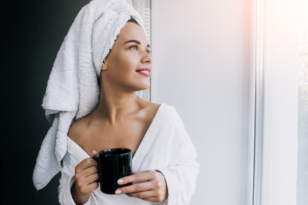 Porträt der jungen schönen Frau im weißen Handtuch und im Gewand, die Kaffee trinken und nahe am Fenster zu Hause genießen