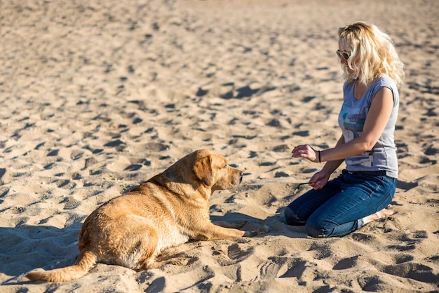 Porträt der jungen schönen Frau in der Sonnenbrille, die auf Sandstrand mit goldenem Apportierhundhund sitzt. Mädchen mit Hund auf dem Seeweg. Glück und Freundschaft. Haustier und Frau. Frau spielt mit Hund am Meeresufer