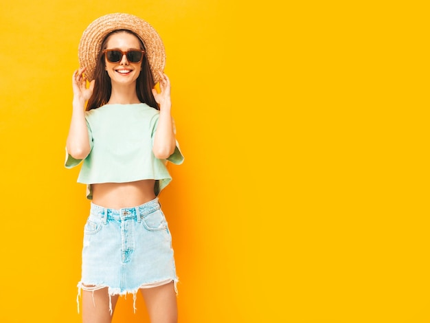 Porträt der jungen schönen lächelnden Frau im trendigen Sommerjeansrock sorglose Frau, die in der Nähe der gelben Wand im Studio posiert Positives Modell, das sich drinnen mit Sonnenbrille amüsiert Fröhlich und glücklich In Hut