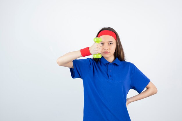 Porträt der jungen sportlichen Frau im blauen T-Shirt, das Hantel hält. muñequera roja