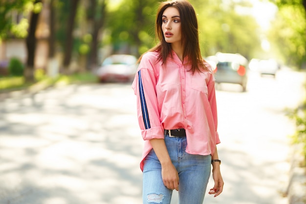 Porträt der jungen und schönen Frau in der Freizeitkleidung auf der Straße. gekleidet in ein rosa Hemd und Jeans. Frühjahr / Sommer-Konzept. Zeit zum Entspannen. Mädchen mit blauen Augen. Genießen Sie den Sonnenuntergang auf der Straße